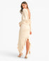 A woman modeling a long-sleeve cream maxi dress with a draped open back and ruffled hem, shown from the back against a plain white studio background.