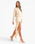 A woman modeling a light beige two-piece outfit consisting of a long-sleeve top and a matching ruffled high-slit skirt, paired with beige strappy high-heeled sandals, against a solid white studio background.