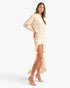 A woman modeling a cream-colored two-piece outfit consisting of a long-sleeve top and a ruffled high-slit skirt, paired with clear-strap high-heeled sandals, against a solid white studio background.