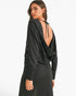 A woman modeling a black long-sleeve dress from the back, showing a draped open back with tie details, on a plain white studio background.