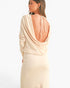 A woman shown from the back wearing a draped, long-sleeve maxi dress in a light cream or beige color. The dress features a deep open back with tie straps and a gathered, draped neckline.