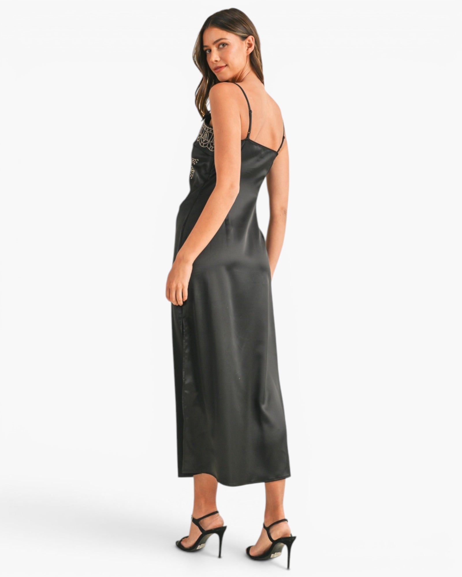 A woman modeling a black satin slip dress with spaghetti straps and a subtle embellished bodice, shown from a three-quarters back angle against a plain white studio background.