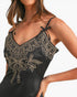 A close-up of a model wearing a black satin camisole slip with beige embroidery and tie straps.