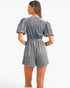 A woman shown from the back wearing a gray velvet short romper with puff sleeves and an elastic waist, photographed against a plain white studio background.