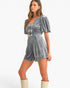 A woman modeling a short gray velvet romper with puff sleeves, a deep V-neck, and decorative bow details down the front, paired with cream knee-high boots against a plain white studio background.