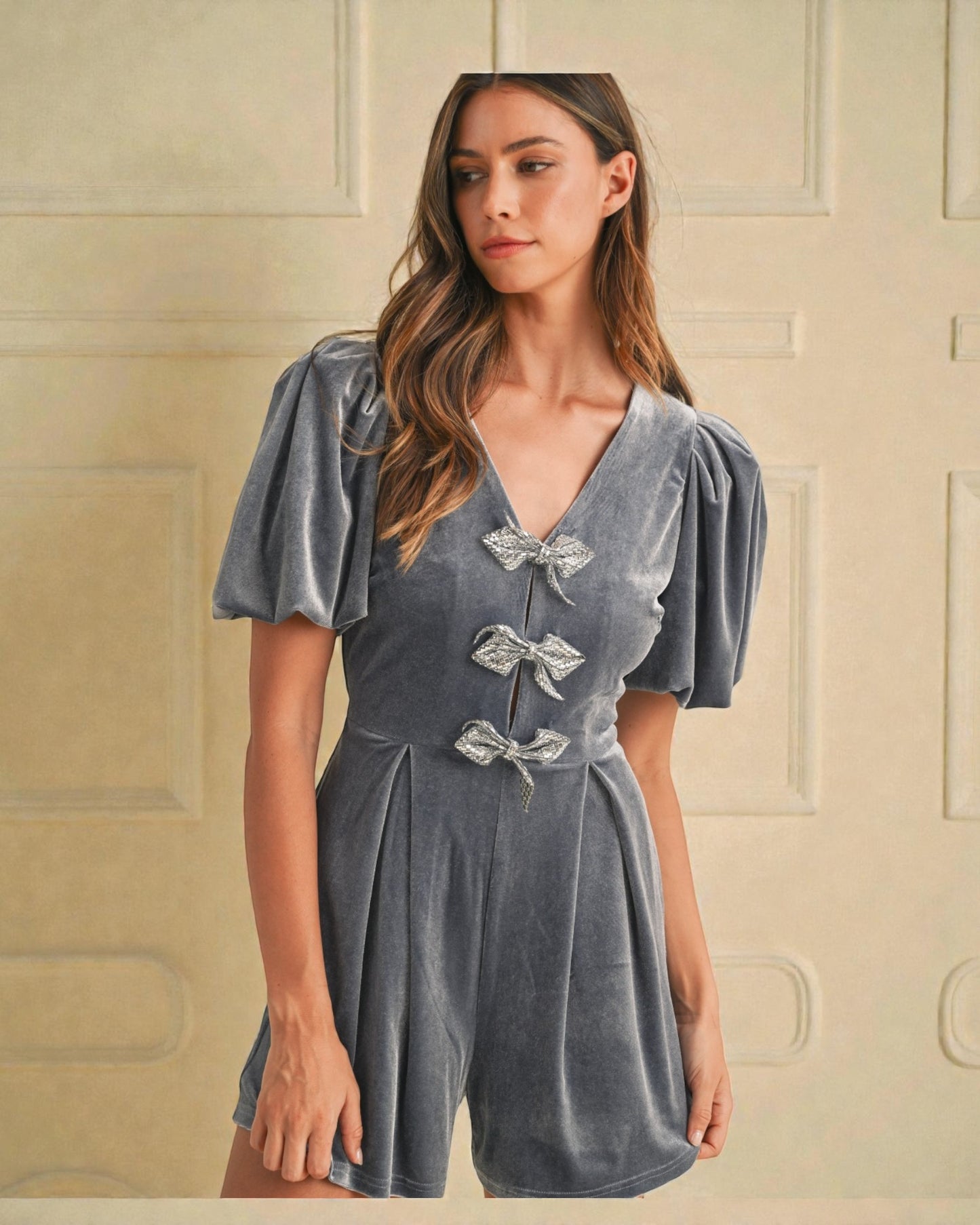 Woman wearing a gray velvet romper with puff sleeves against a beige wall.