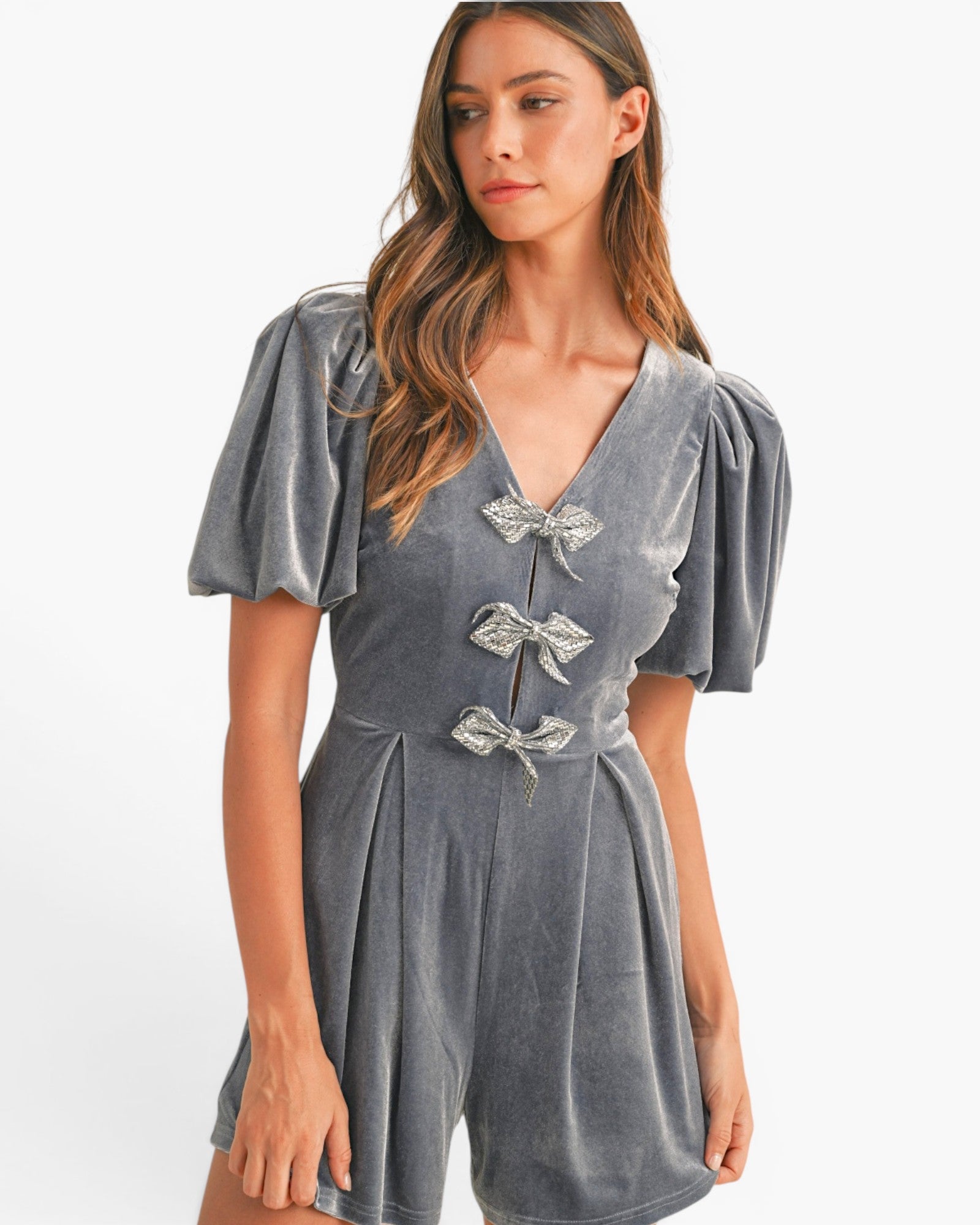 A woman wearing a gray velvet short-sleeve romper with puff sleeves, a V-neckline, and three decorative bow details on the front, set against a plain white studio background.