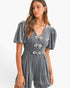 A woman wearing a gray velvet short-sleeve romper with puff sleeves, a V-neckline, and three decorative bow details on the front, set against a plain white studio background.