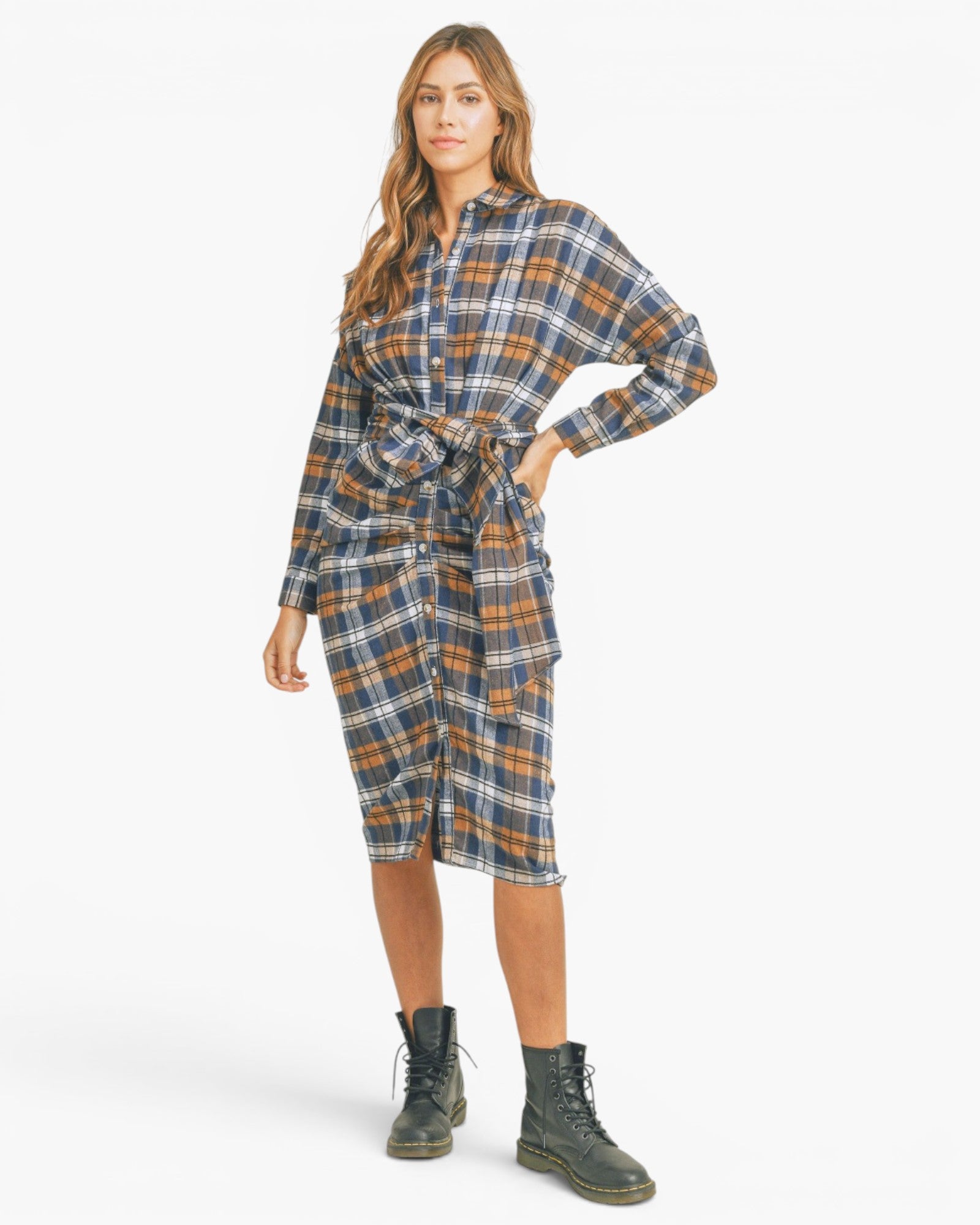 A woman modeling a long-sleeve plaid shirt dress with a tie waist, shown against a plain light gray studio background. She is wearing dark green lace-up boots.