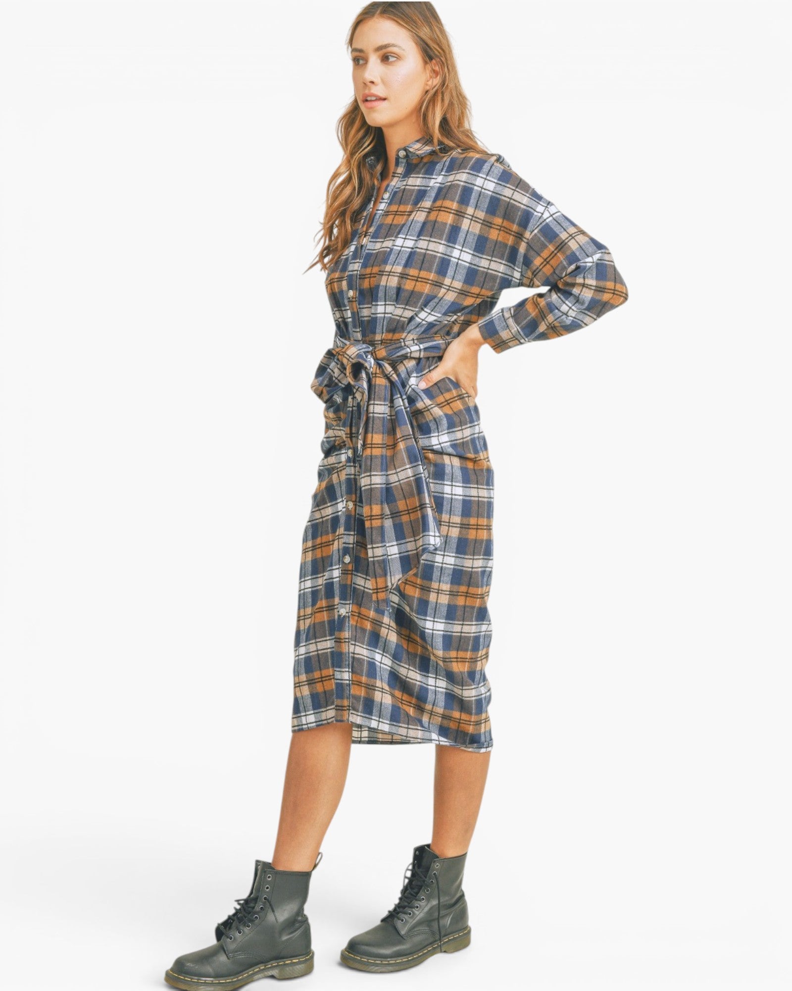 A woman modeling a long-sleeve plaid shirt dress with a tie waist, shown in profile against a plain white studio background. She is wearing dark green combat boots.