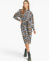 A woman modeling a long-sleeve plaid shirt dress with a tie waist, shown against a plain light gray studio background. The dress has a blue, brown, and white plaid pattern, and she is wearing black lace-up boots.