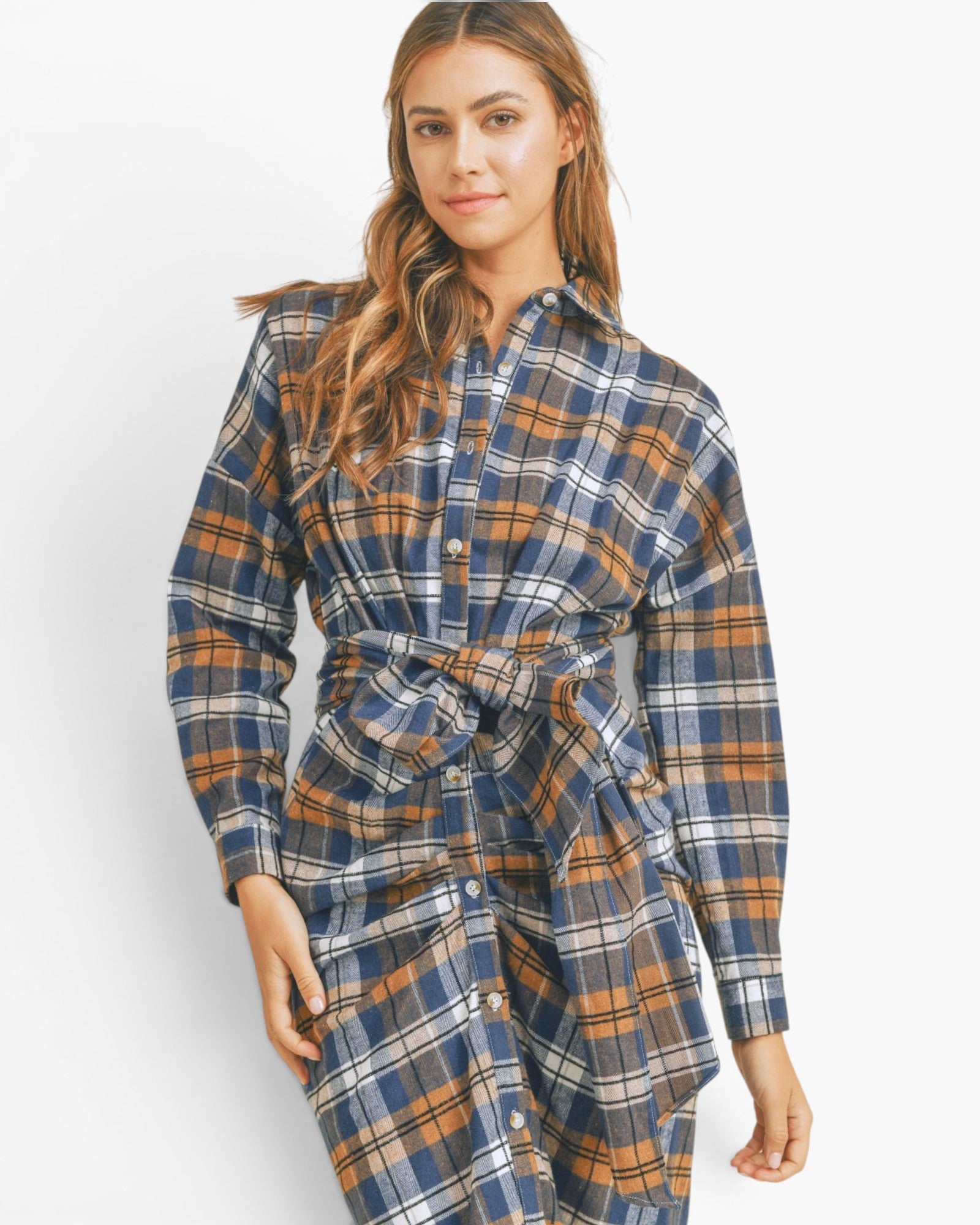 A woman modeling a long-sleeve plaid shirt dress in navy blue, orange, and white. The dress features a button-down front, a collar, and a self-tie belt at the waist, set against a plain white studio background.