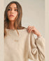 Woman wearing a beige sweatshirt with a brand logo, holding it in front of a neutral background