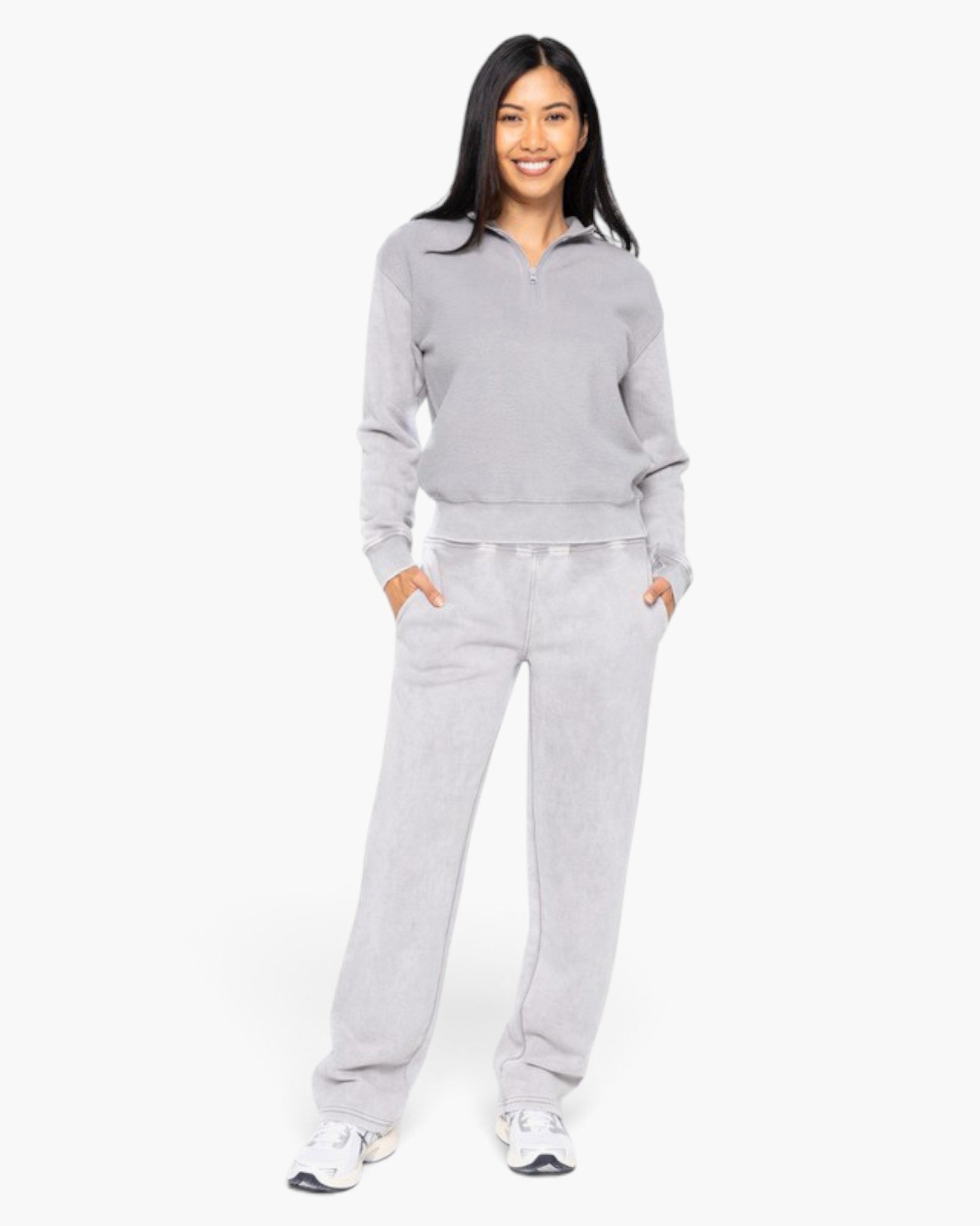 A woman modeling a light gray two-piece loungewear set consisting of a quarter-zip sweatshirt and matching sweatpants, standing against a plain white studio background.