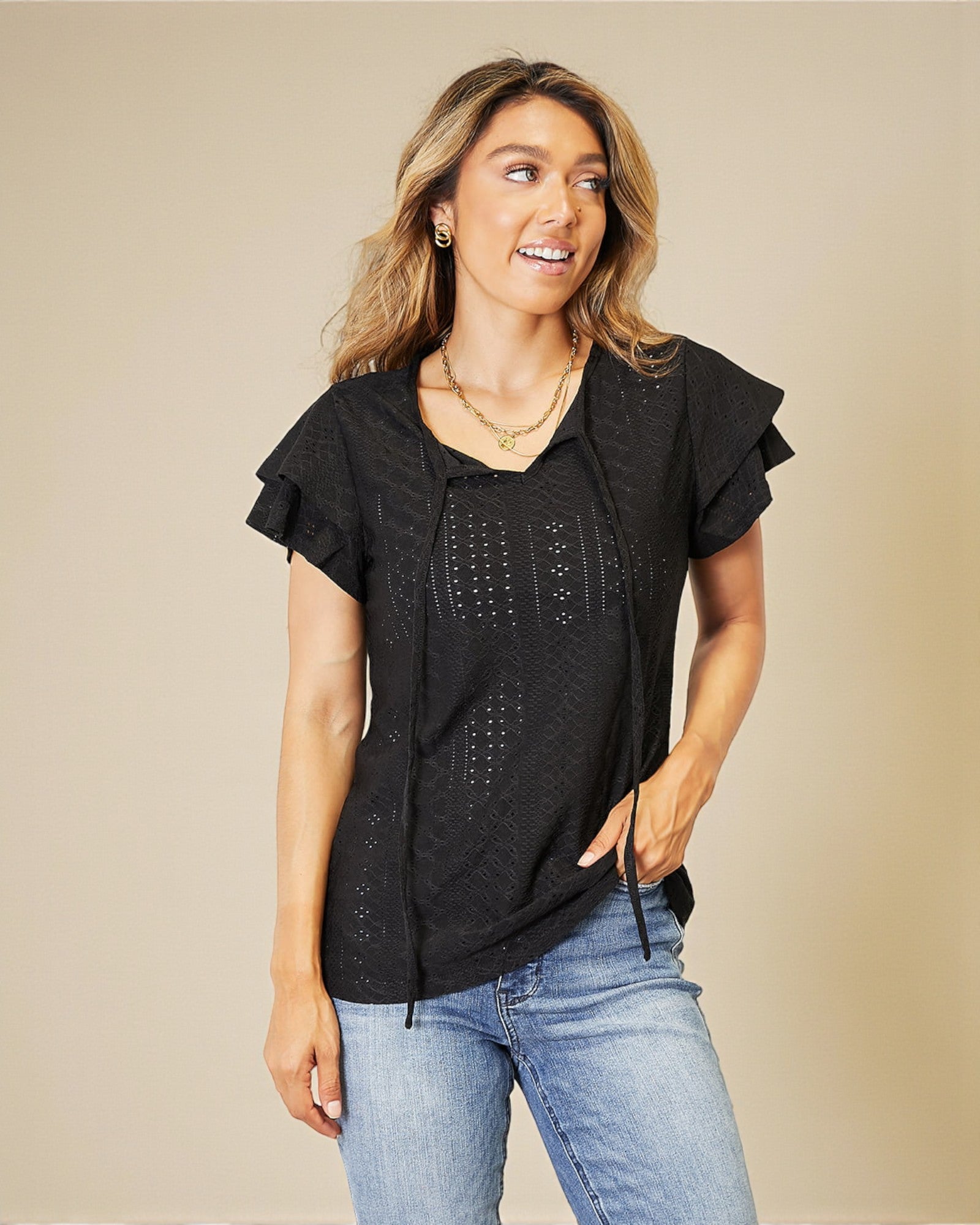 Woman wearing a black top with ruffled sleeves and blue jeans on a beige background