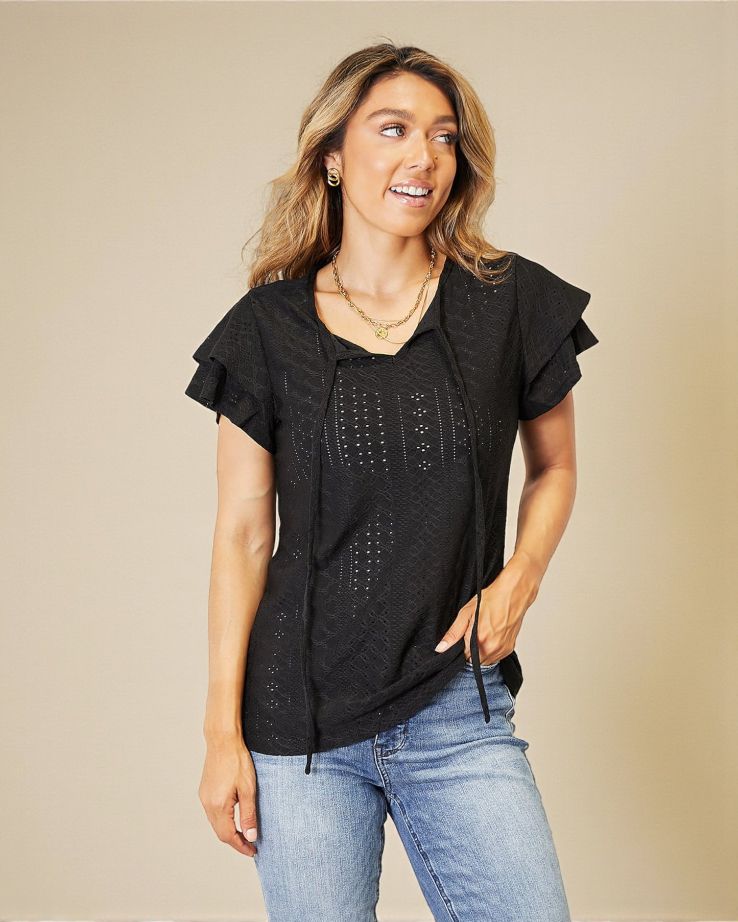 Woman wearing a black top with ruffled sleeves and blue jeans on a beige background