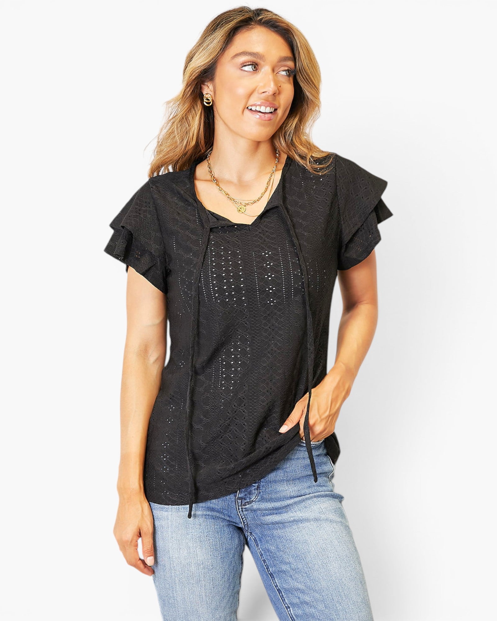 A woman wearing a black short-sleeve blouse with ruffled sleeves and a tie-front detail, paired with light blue jeans, photographed against a plain light gray background.