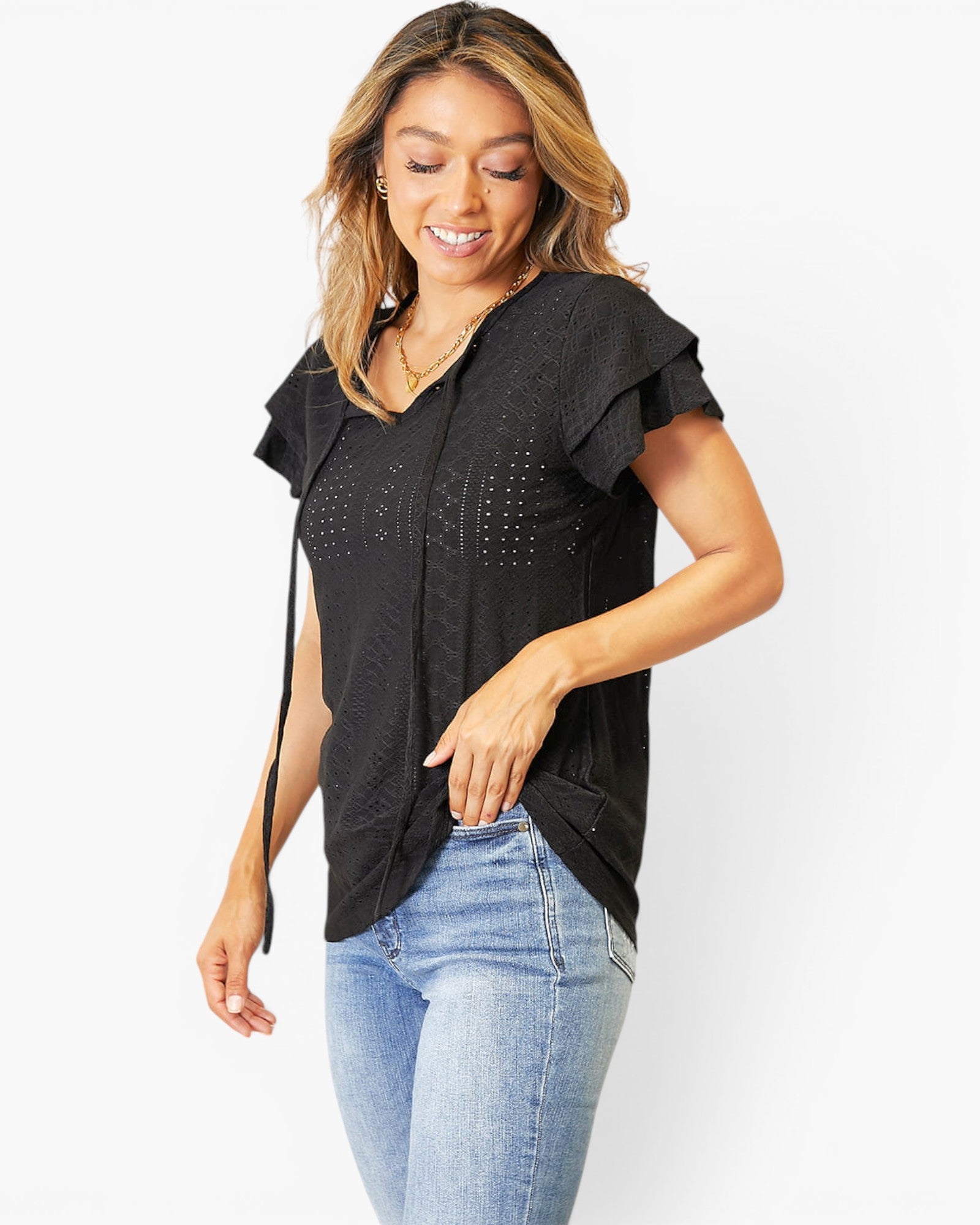 A woman wearing a black short-sleeve top with ruffle details and blue jeans, standing against a plain white background.