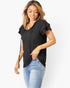 A woman wearing a black short-sleeve top with ruffle details and blue jeans, standing against a plain white background.