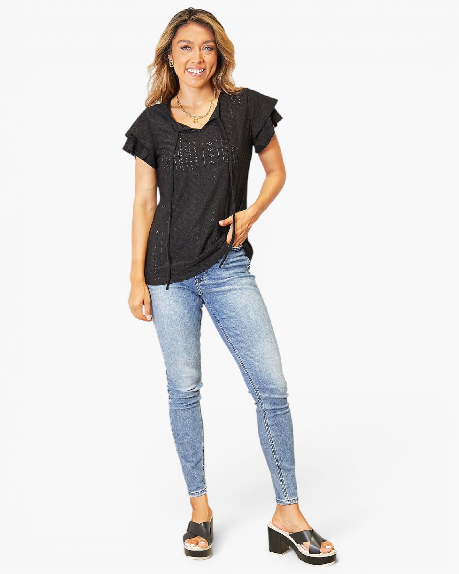 A woman modeling a black short-sleeve top with ruffled sleeves and decorative eyelet embroidery on the chest, paired with light-wash blue skinny jeans and black slide sandals, against a plain white background.