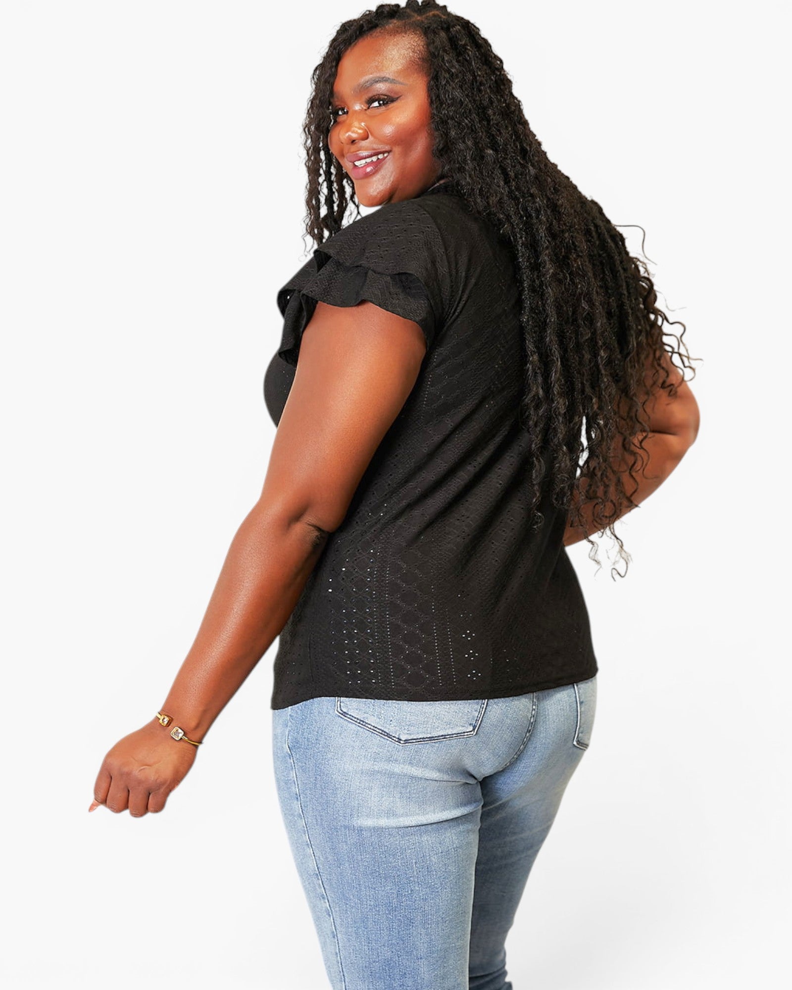 A woman with long black dreadlocks is shown from a side-back angle wearing a black short-sleeve top with ruffled sleeves and light-wash blue jeans, against a plain white studio background.