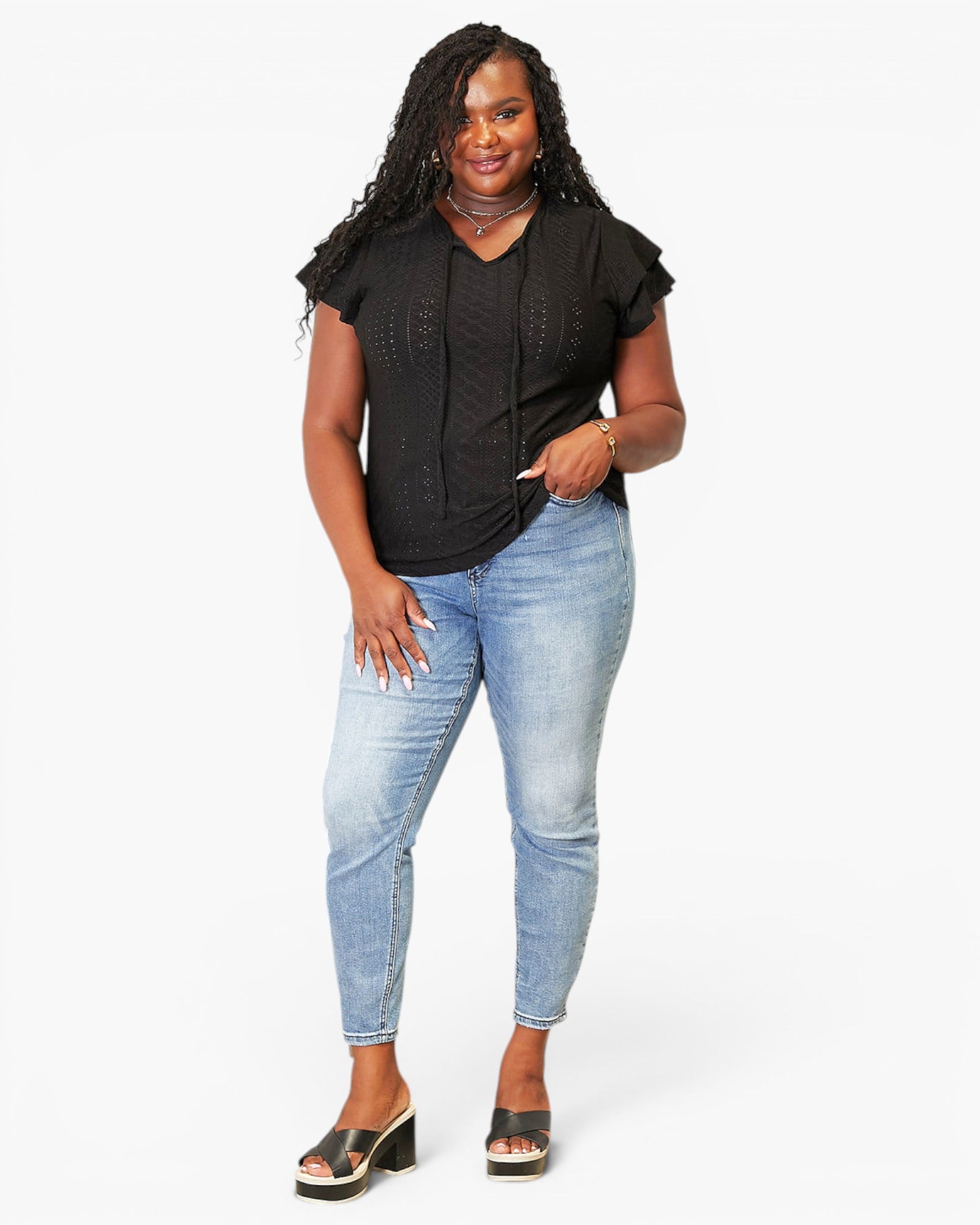 A woman modeling a black short-sleeve top with lace-up front detailing, paired with light-wash blue skinny jeans and black slide sandals, against a plain white studio background.