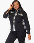 A woman modeling a black button-front shirt jacket with plaid trim details on the collar, placket, chest pocket, and side seams, paired with black pants against a light gray studio background.