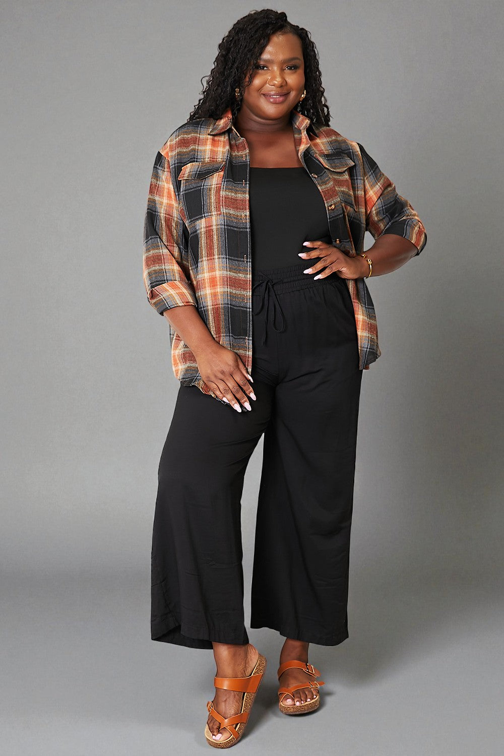 Woman wearing a plaid shirt over a black top and wide-leg pants on a gray background