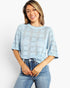 A woman wearing a light blue knit top with a geometric openwork pattern and blue jeans, standing against a plain light gray background.