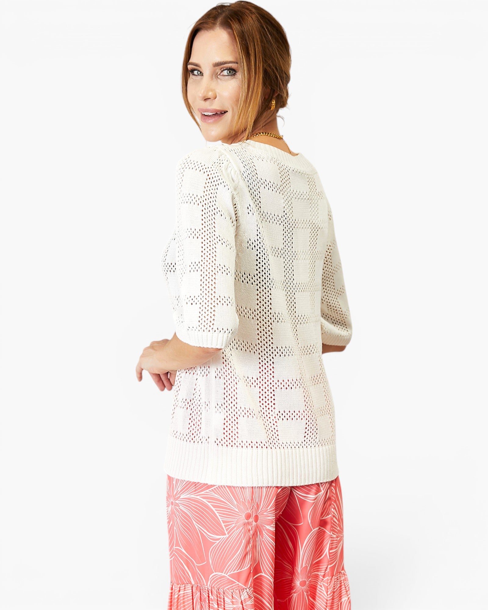 A woman wearing a cream-colored knit sweater with a geometric openwork pattern and pink floral print pants, photographed against a plain white studio background.