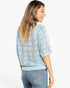 A woman shown from the back wearing a light blue openwork knit sweater with a geometric pattern and blue jeans, against a plain white studio background.