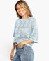A woman wearing a light blue open-knit sweater with a geometric pattern and blue jeans, posing against a plain white studio background.