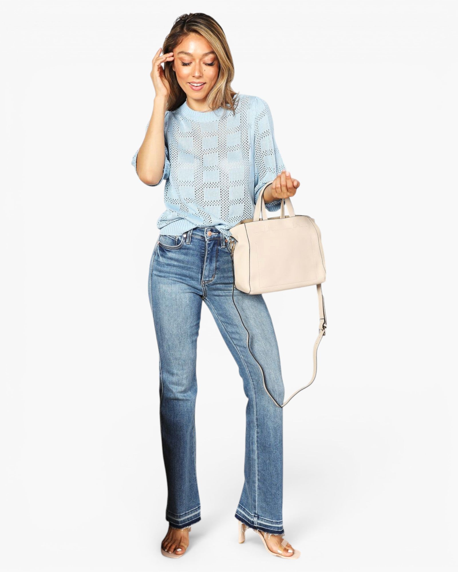 A woman modeling a light blue patterned top, blue jeans, and nude heels while holding a light beige handbag against a plain white background.