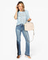 A woman modeling a light blue patterned top, blue jeans, and nude heels while holding a light beige handbag against a plain white background.