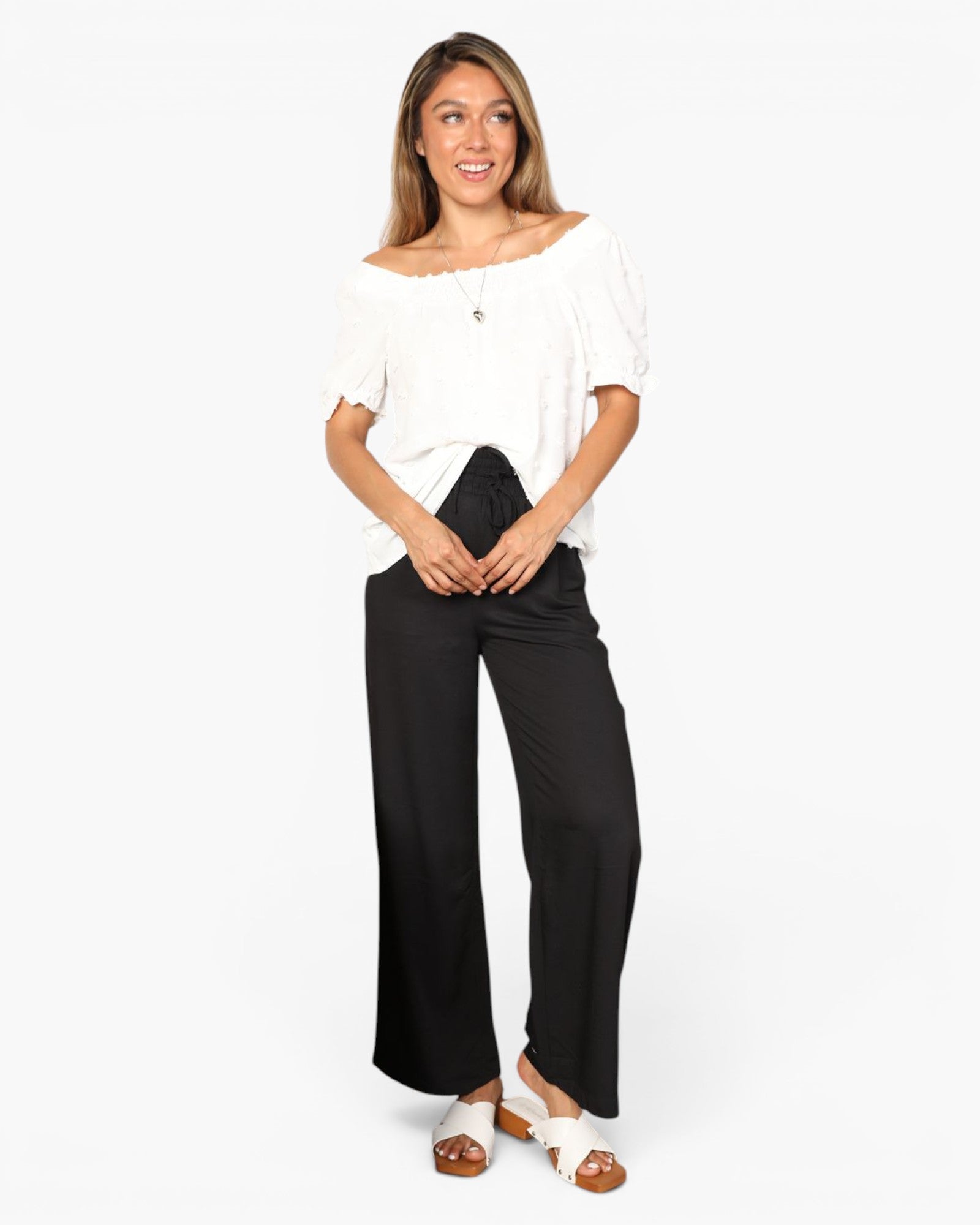A woman modeling an off-the-shoulder white puff-sleeve top and black wide-leg pants, paired with white slide sandals, against a plain white studio background.