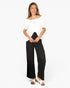 A woman modeling an off-the-shoulder white puff-sleeve top and black wide-leg pants, paired with white slide sandals, against a plain white studio background.