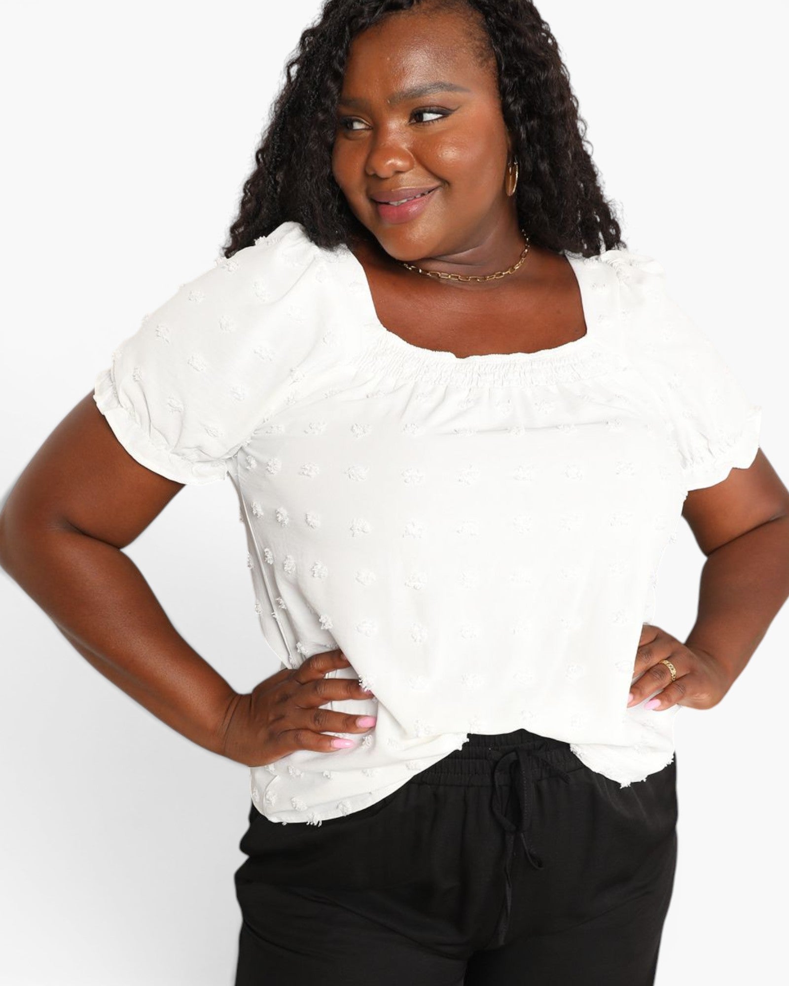 A woman wearing a white short-sleeve top with a textured, dotted fabric and a square neckline, paired with black pants, against a plain white background.