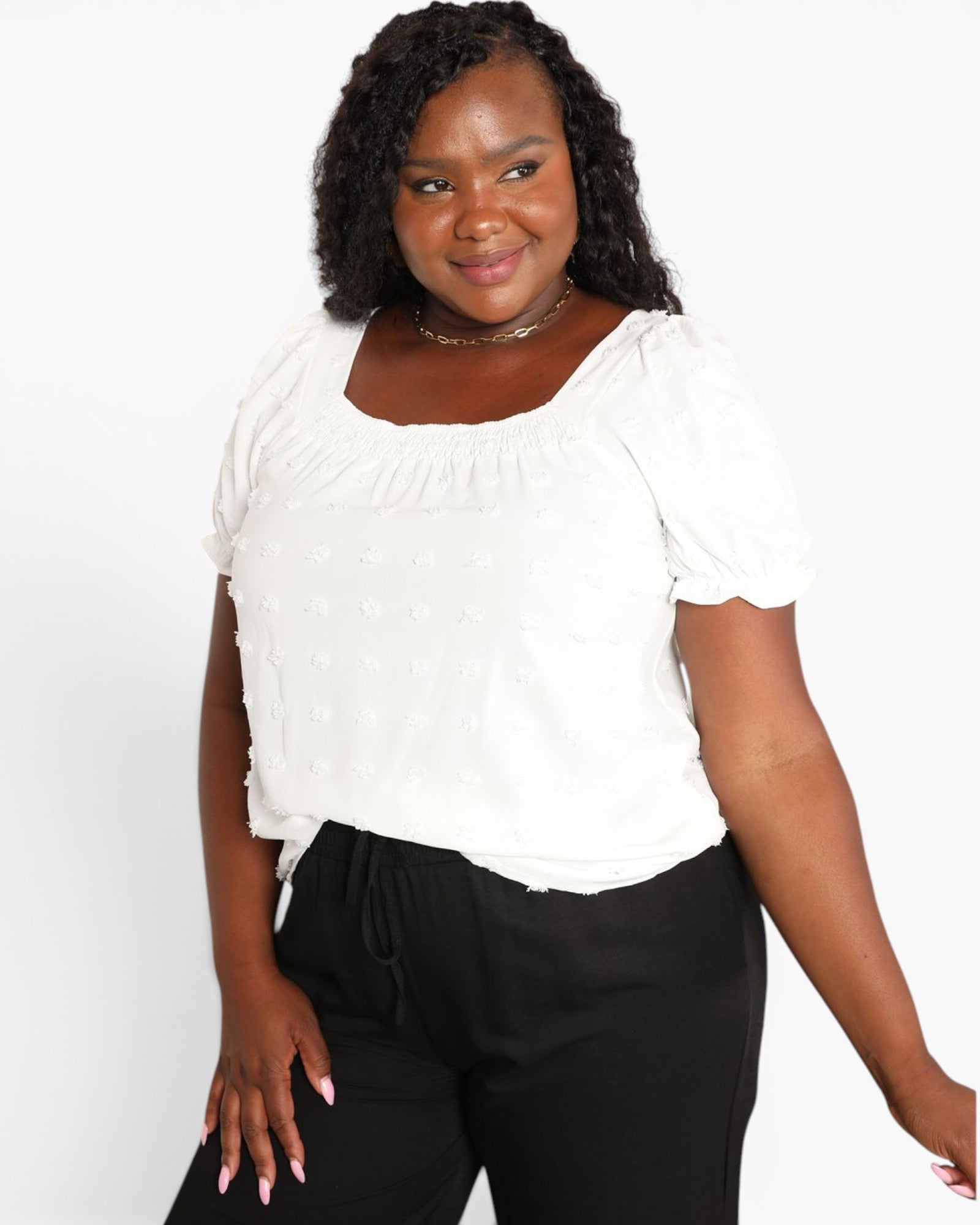 A woman wearing a white short-sleeve puff-sleeve top with a square neckline and black pants, standing against a light gray studio background.