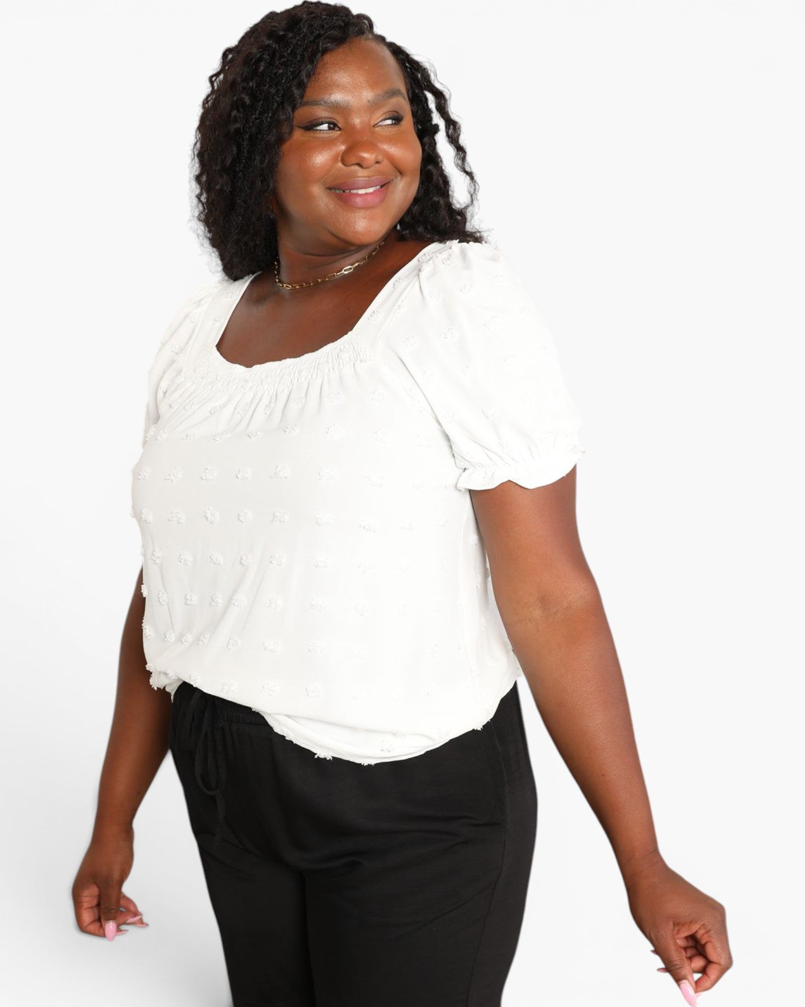 A woman wearing a white short-sleeve top with a gathered neckline and black pants, photographed against a light gray studio background.