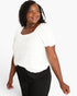 A woman wearing a white short-sleeve top with a gathered neckline and black pants, photographed against a light gray studio background.