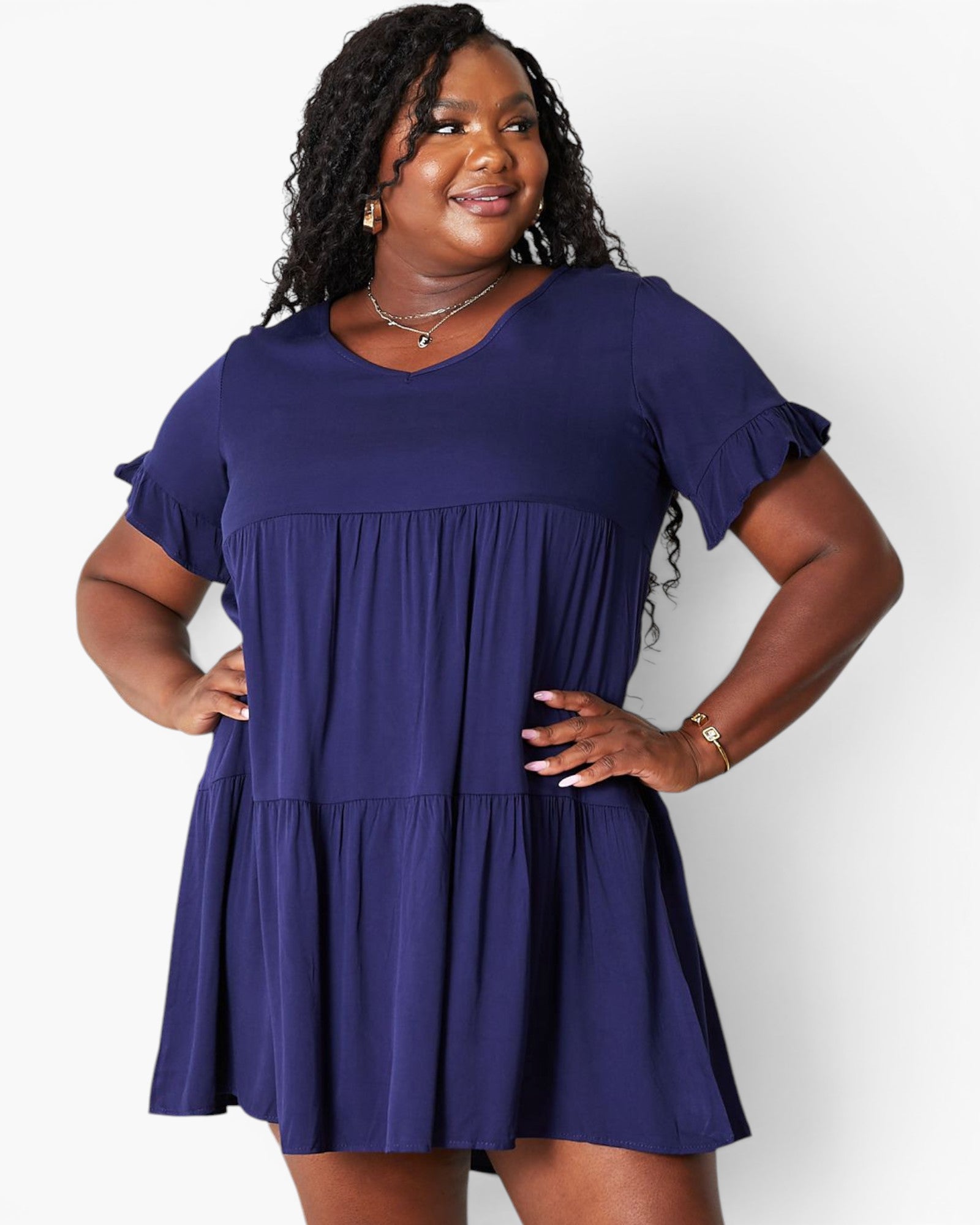 A woman modeling a short-sleeve tiered mini dress in a deep purple color, standing against a light gray studio background.