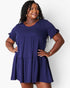 A woman modeling a short-sleeve tiered mini dress in a deep purple color, standing against a light gray studio background.