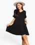 A woman wearing a black tiered short-sleeve dress with ruffled sleeves and side pockets, paired with a light beige wide-brim hat, posing against a solid white background.