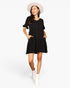 A woman modeling a black short-sleeve mini dress with ruffled sleeves and front patch pockets, styled with a light beige wide-brim hat and white ankle boots against a plain white studio background.