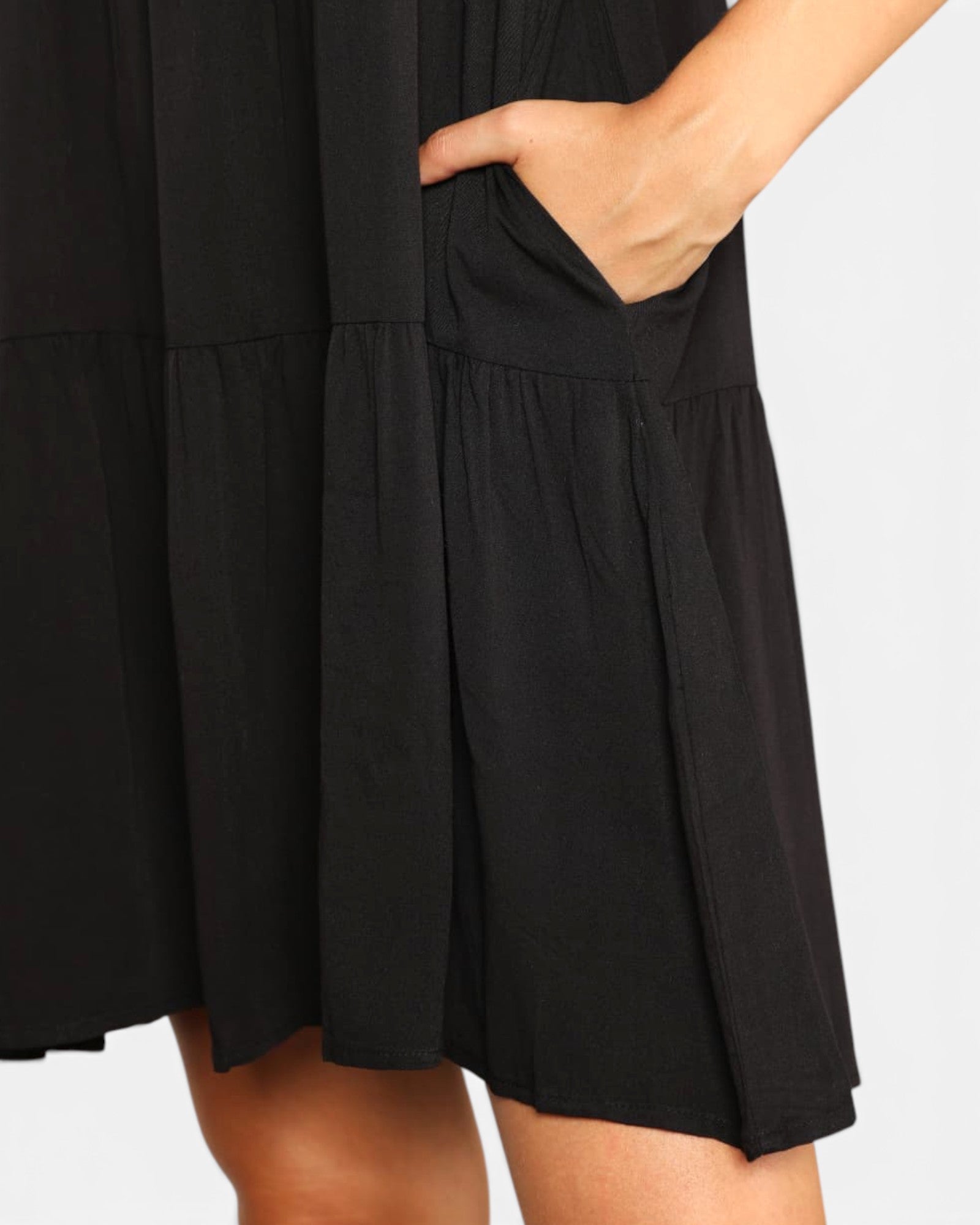Close-up of a black tiered dress with a side pocket, shown on a model with one hand in the pocket against a light gray background.