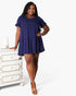 A woman modeling a short-sleeve navy blue tiered mini dress, standing next to a white dresser against a light gray studio background.