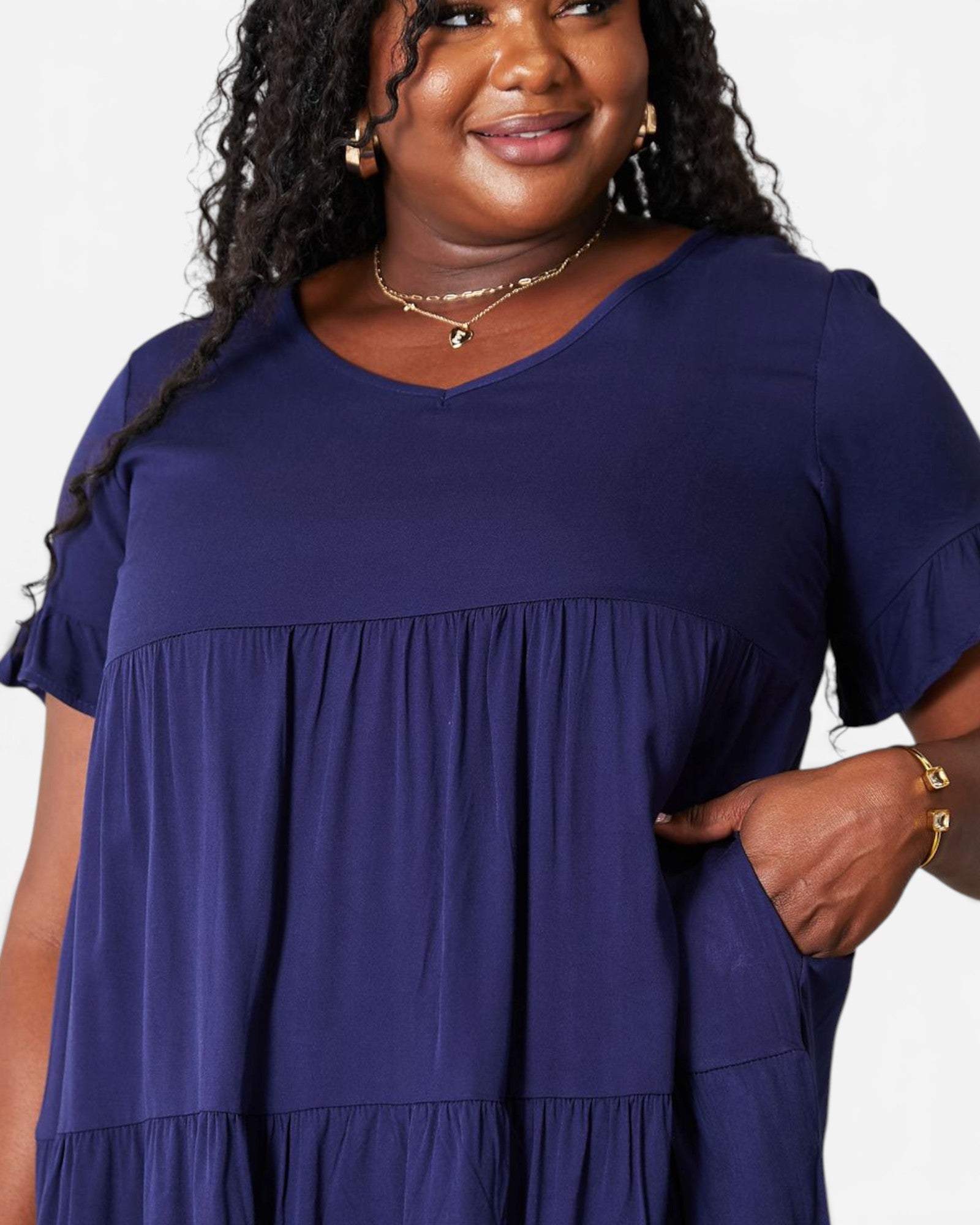 A woman wearing a navy blue tiered short-sleeve tunic dress with ruffled sleeves and side pockets, shown from the chest up against a plain light background.