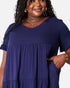 A woman wearing a navy blue tiered short-sleeve tunic dress with ruffled sleeves and side pockets, shown from the chest up against a plain light background.
