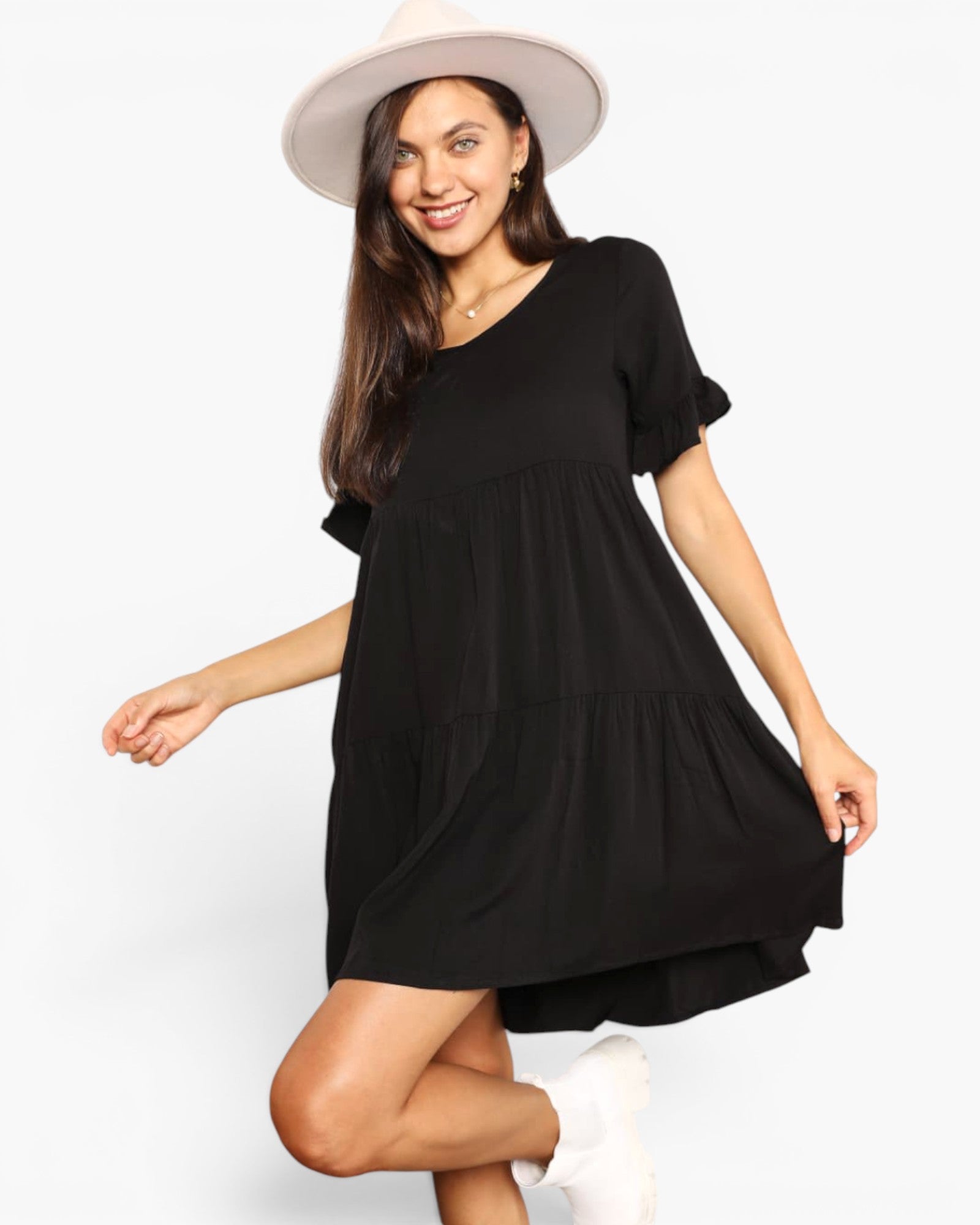 A woman modeling a black tiered mini dress with short flutter sleeves and a V-neck, paired with a wide-brimmed light beige hat and white sneakers against a plain light gray studio background.