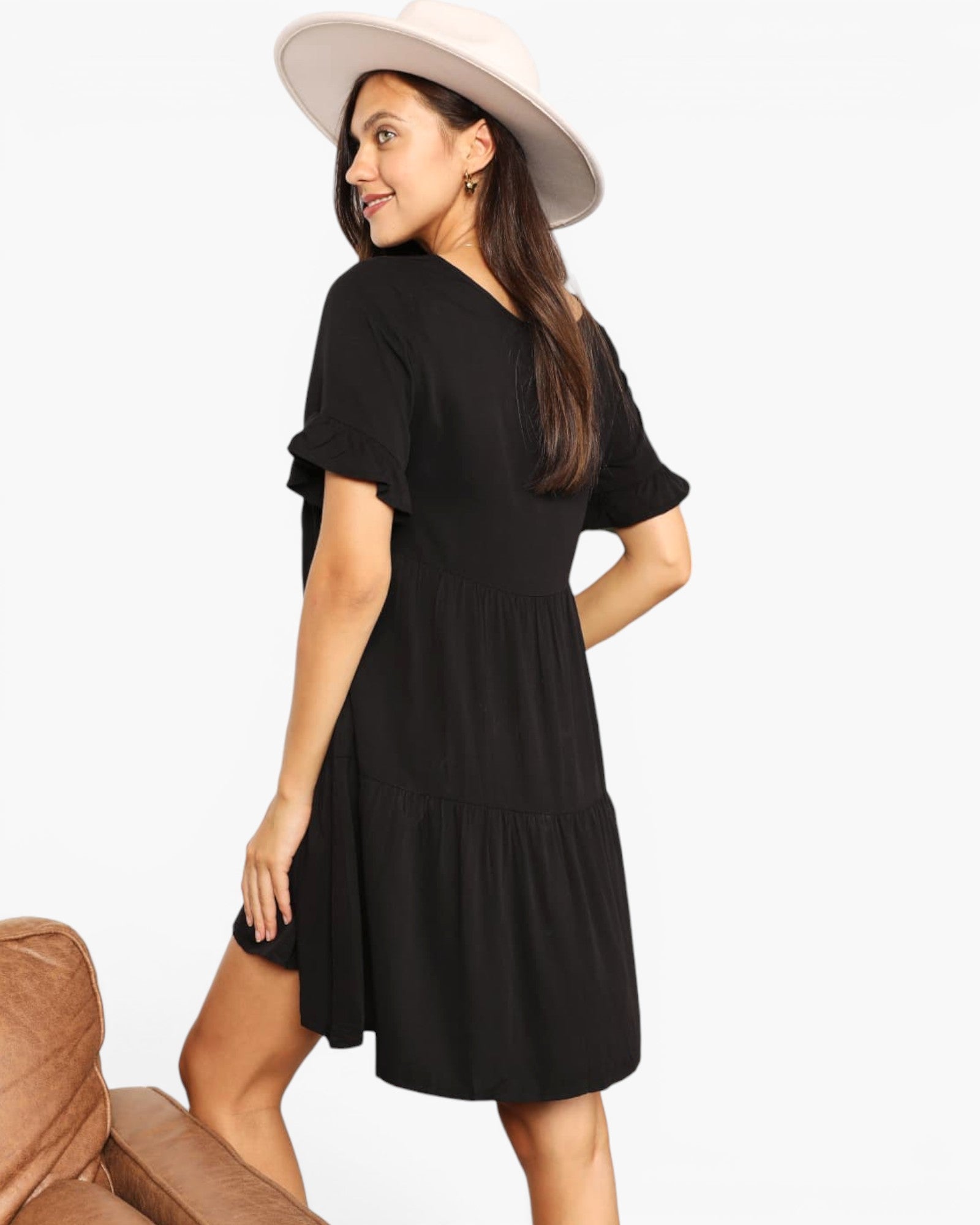 A woman wearing a black tiered short-sleeve dress and a wide-brimmed cream hat, photographed against a plain light background with part of a brown chair visible at the lower left.